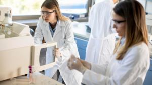 Pesquisadoras usando vestimenta de proteção estão mexendo em equipamentos de cromatografia