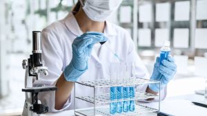 cientista com tubos de ensaio e pipeta, usando jaleco brando, máscara de luvas azuis